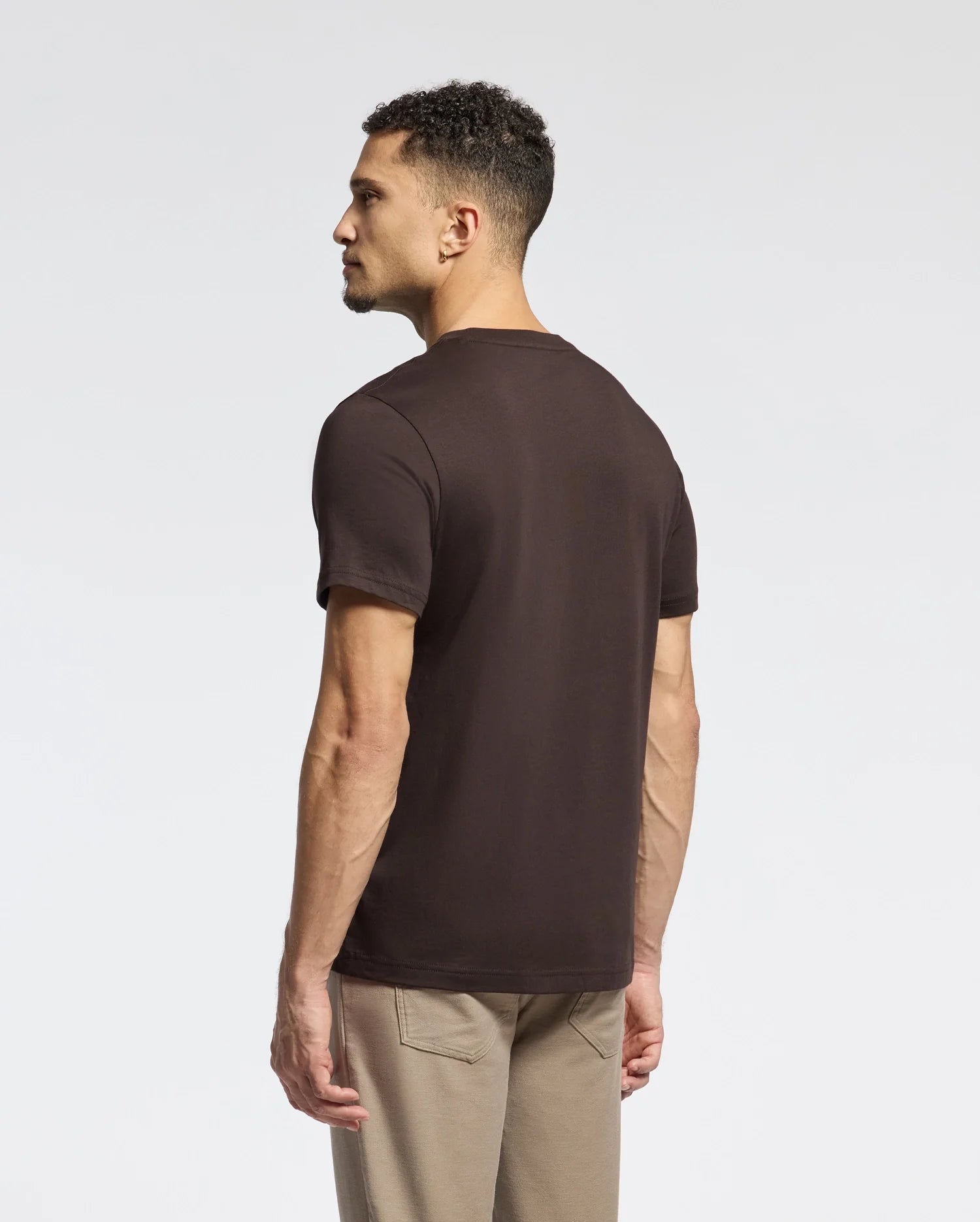 A man with short curly hair stands slightly to the side, wearing a Psycho Bunny Men's Monarc Front Bunny Graphic Tee (B6U211E200) made from soft Pima cotton, paired with light-colored pants against a plain white background.