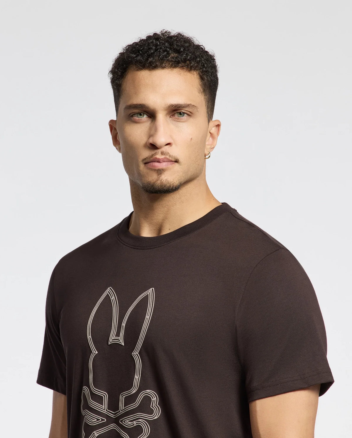 A man with short curly hair is wearing a Psycho Bunny Men's Monarc Front Bunny Graphic Tee, crafted from soft Pima cotton. The black t-shirt features a raised-printed bunny with crossbones design against a plain, light-colored background.