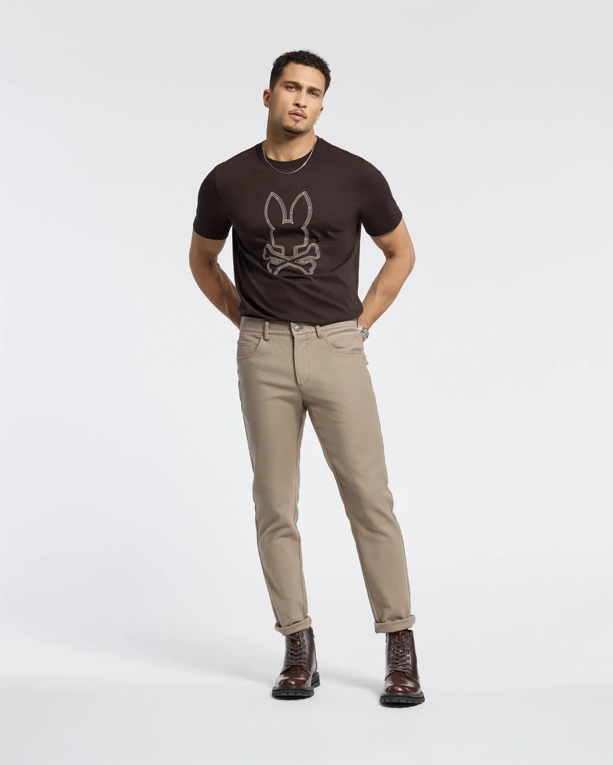 A man stands against a plain white background, wearing a black MENS MONARC FRONT BUNNY GRAPHIC TEE - B6U211E200 by Psycho Bunny, paired with beige pants and dark brown boots. With his hands in his pockets and a direct gaze at the camera, he epitomizes effortless style.