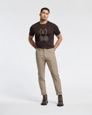 A man stands against a plain white background, wearing a black MENS MONARC FRONT BUNNY GRAPHIC TEE - B6U211E200 by Psycho Bunny, paired with beige pants and dark brown boots. With his hands in his pockets and a direct gaze at the camera, he epitomizes effortless style.