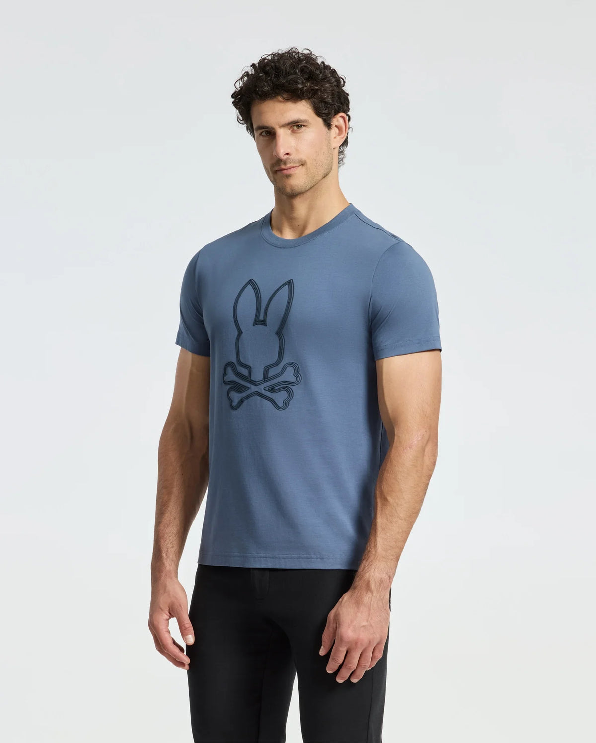 Against a plain white backdrop, an individual dons the MENS MONARC FRONT BUNNY GRAPHIC TEE - B6U211E200 by Psycho Bunny. Crafted from soft Pima cotton, this blue tee showcases a stylized rabbit above crossed bones. With curly dark hair and black pants, they project a cool and fashionable aura.