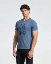 Against a plain white backdrop, an individual dons the MENS MONARC FRONT BUNNY GRAPHIC TEE - B6U211E200 by Psycho Bunny. Crafted from soft Pima cotton, this blue tee showcases a stylized rabbit above crossed bones. With curly dark hair and black pants, they project a cool and fashionable aura.