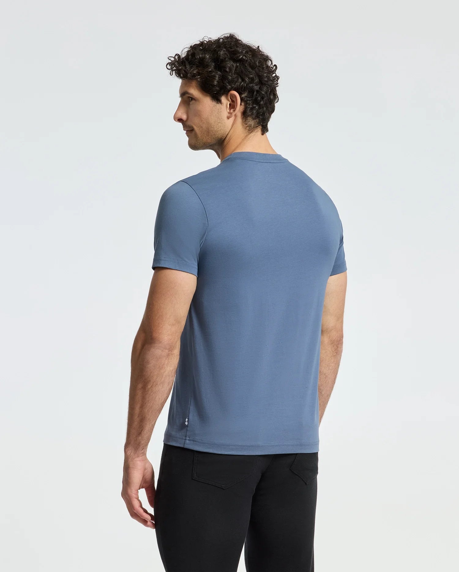 A person with curly hair is standing sideways, wearing the MENS MONARC FRONT BUNNY GRAPHIC TEE - B6U211E200 by Psycho Bunny, a blue Pima cotton short-sleeve t-shirt showcasing a playful bunny graphic, paired with black pants against a plain background.