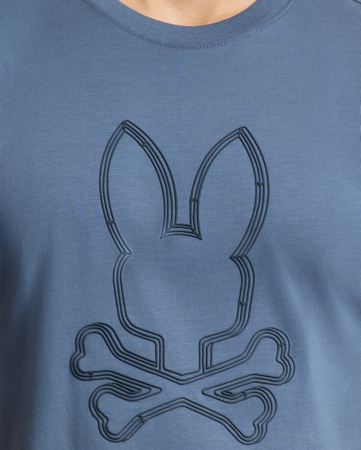 A close-up image of the MENS MONARC FRONT BUNNY GRAPHIC TEE - B6U211E200 from Psycho Bunny, made from soft Pima cotton. It showcases a raised bunny silhouette design above two crossed bone shapes, similar to a skull and crossbones theme.