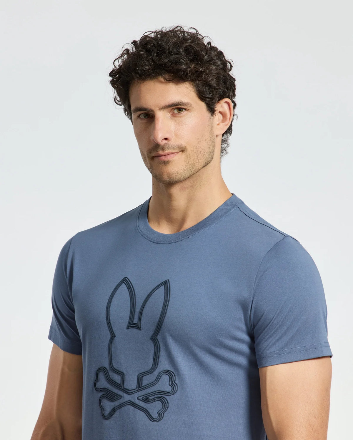 A man is wearing the Psycho Bunny MENS MONARC FRONT BUNNY GRAPHIC TEE - B6U211E200, made from soft Pima cotton and featuring a stylized bunny logo with crossed bones below. The design stands out against a plain, light background, highlighting its Monarc-inspired appeal.