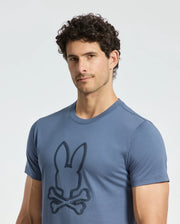 A man is wearing the Psycho Bunny MENS MONARC FRONT BUNNY GRAPHIC TEE - B6U211E200, made from soft Pima cotton and featuring a stylized bunny logo with crossed bones below. The design stands out against a plain, light background, highlighting its Monarc-inspired appeal.