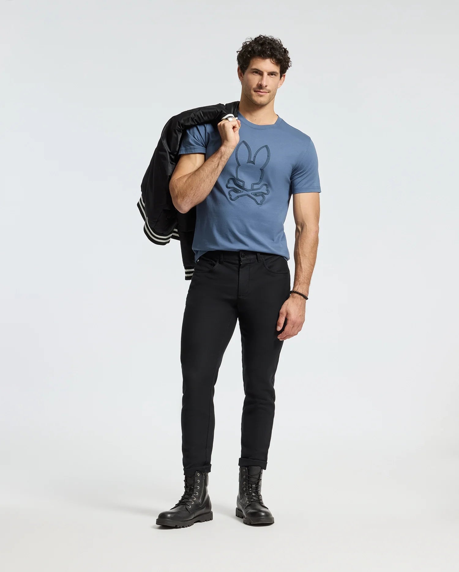 A man with curly hair stands confidently, holding a jacket over his shoulder. He's wearing a Psycho Bunny MENS MONARC FRONT BUNNY GRAPHIC TEE - B6U211E200 made from pima cotton, paired with black fitted pants and black boots against a plain white background.