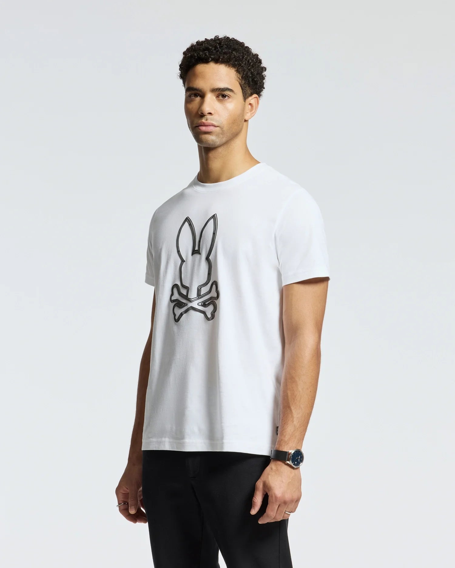A man stands wearing a Psycho Bunny MENS MONARC FRONT BUNNY GRAPHIC TEE - B6U211E200, made from 100% Pima cotton in Peru. The tee prominently displays a logo with a bunny and crossbones design. He has short, curly hair and is paired with black pants and a black watch. The background is plain and light gray.