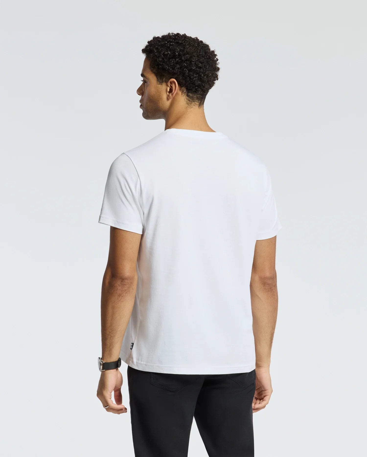 A man with short curly hair is dressed in a minimalist MENS MONARC FRONT BUNNY GRAPHIC TEE - B6U211E200 from Psycho Bunny, crafted from 100% Pima cotton in Peru. He pairs it with black pants as he stands facing away from the camera against a light gray background.