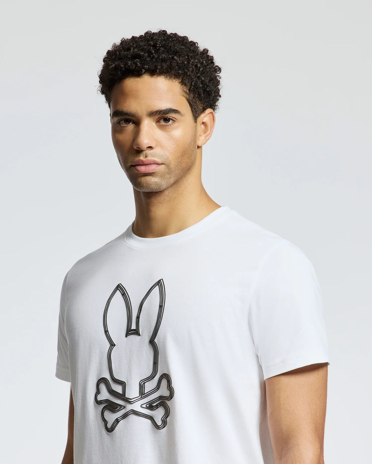 A man with short curly hair is wearing the Psycho Bunny Men's Monarc Front Bunny Graphic Tee, crafted from 100% Pima cotton in Peru. The white T-shirt showcases a bunny with crossbones set against a simple light gray background.