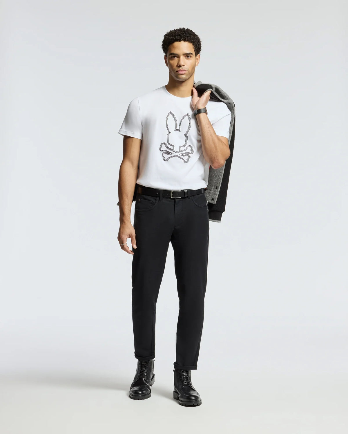 A man stands against a plain, light-colored background with a jacket draped over his shoulder, wearing the Psycho Bunny MENS MONARC FRONT BUNNY GRAPHIC TEE - B6U211E200 in 100% Pima cotton. The shirt features a bunny above crossed bones design. Complemented by black pants and boots, this tee is made in Peru for style and comfort.