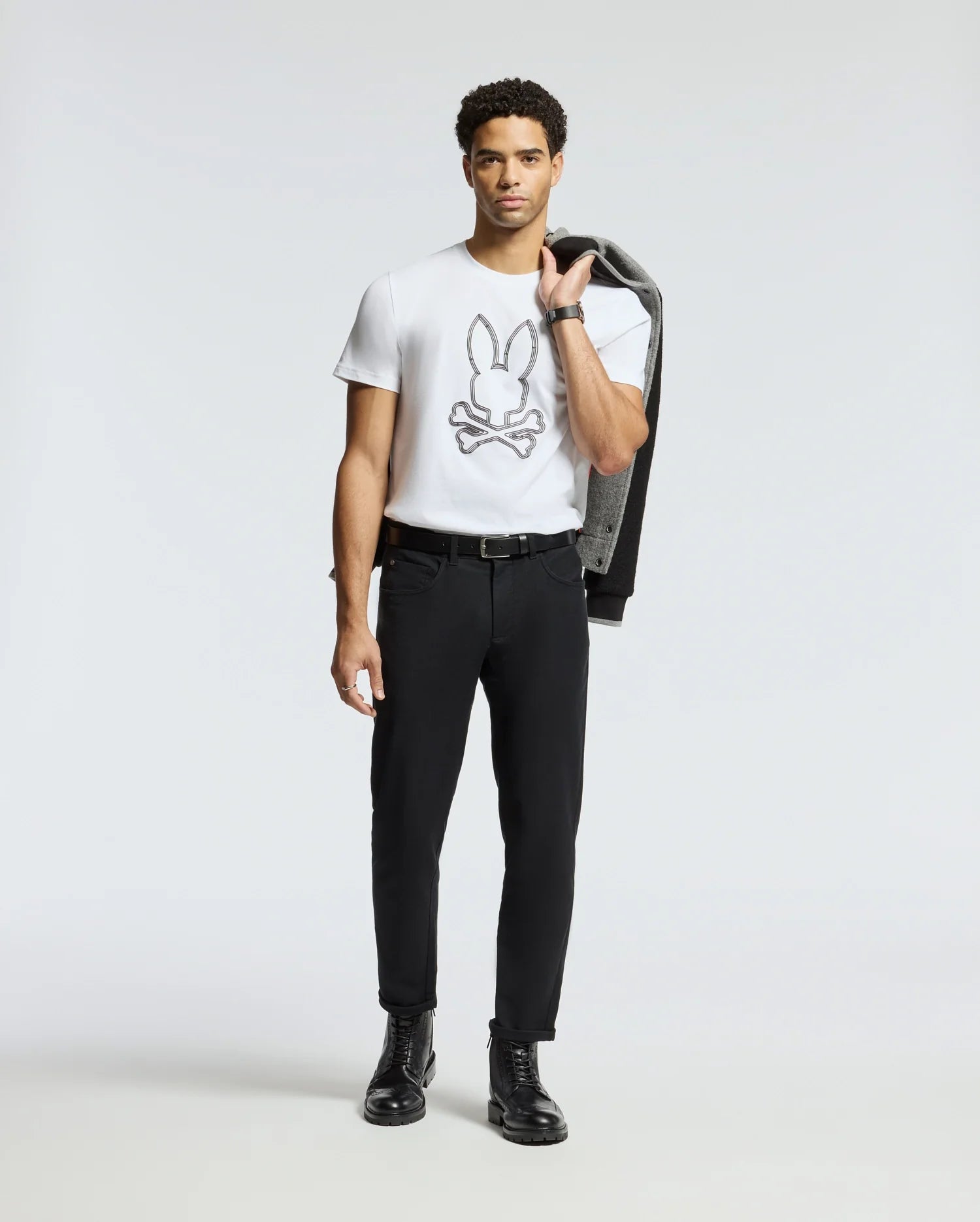 A man stands against a plain, light-colored background with a jacket draped over his shoulder, wearing the Psycho Bunny MENS MONARC FRONT BUNNY GRAPHIC TEE - B6U211E200 in 100% Pima cotton. The shirt features a bunny above crossed bones design. Complemented by black pants and boots, this tee is made in Peru for style and comfort.