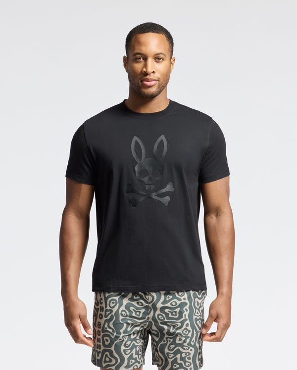 A man is flaunting a Psycho Bunny MENS PAYSON LEATHER PRINT GRAPHIC TEE with a rabbit-eared skull and crossbones design, paired with patterned shorts. The light background accentuates his edgy look.