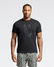 A man is flaunting a Psycho Bunny MENS PAYSON LEATHER PRINT GRAPHIC TEE with a rabbit-eared skull and crossbones design, paired with patterned shorts. The light background accentuates his edgy look.