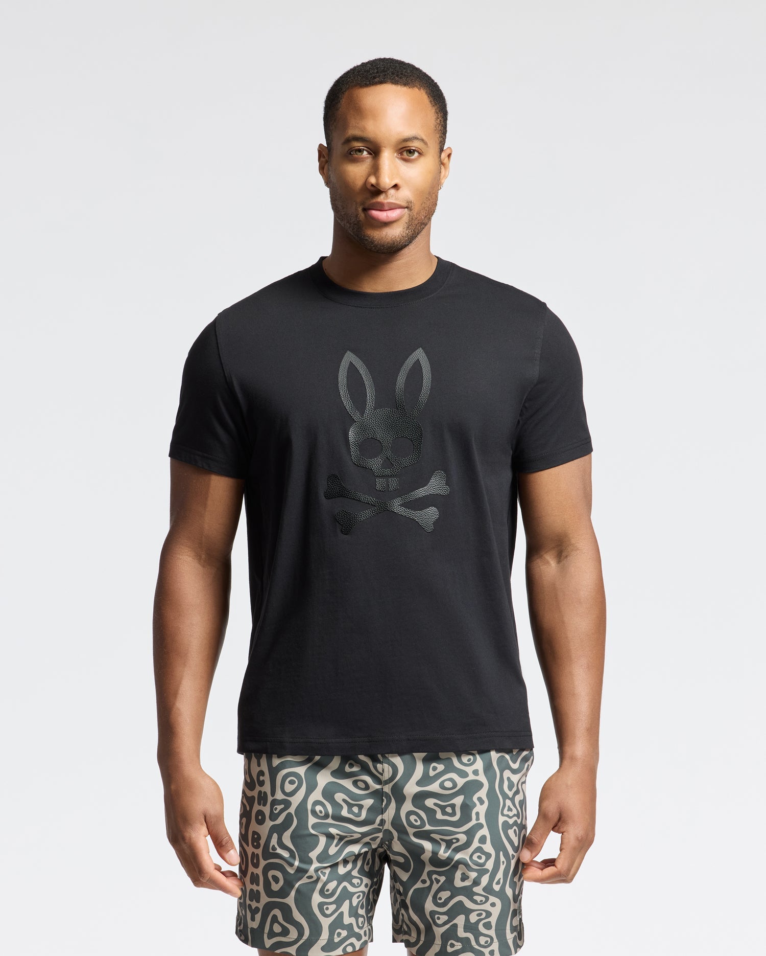 A man is flaunting a Psycho Bunny MENS PAYSON LEATHER PRINT GRAPHIC TEE with a rabbit-eared skull and crossbones design, paired with patterned shorts. The light background accentuates his edgy look.
