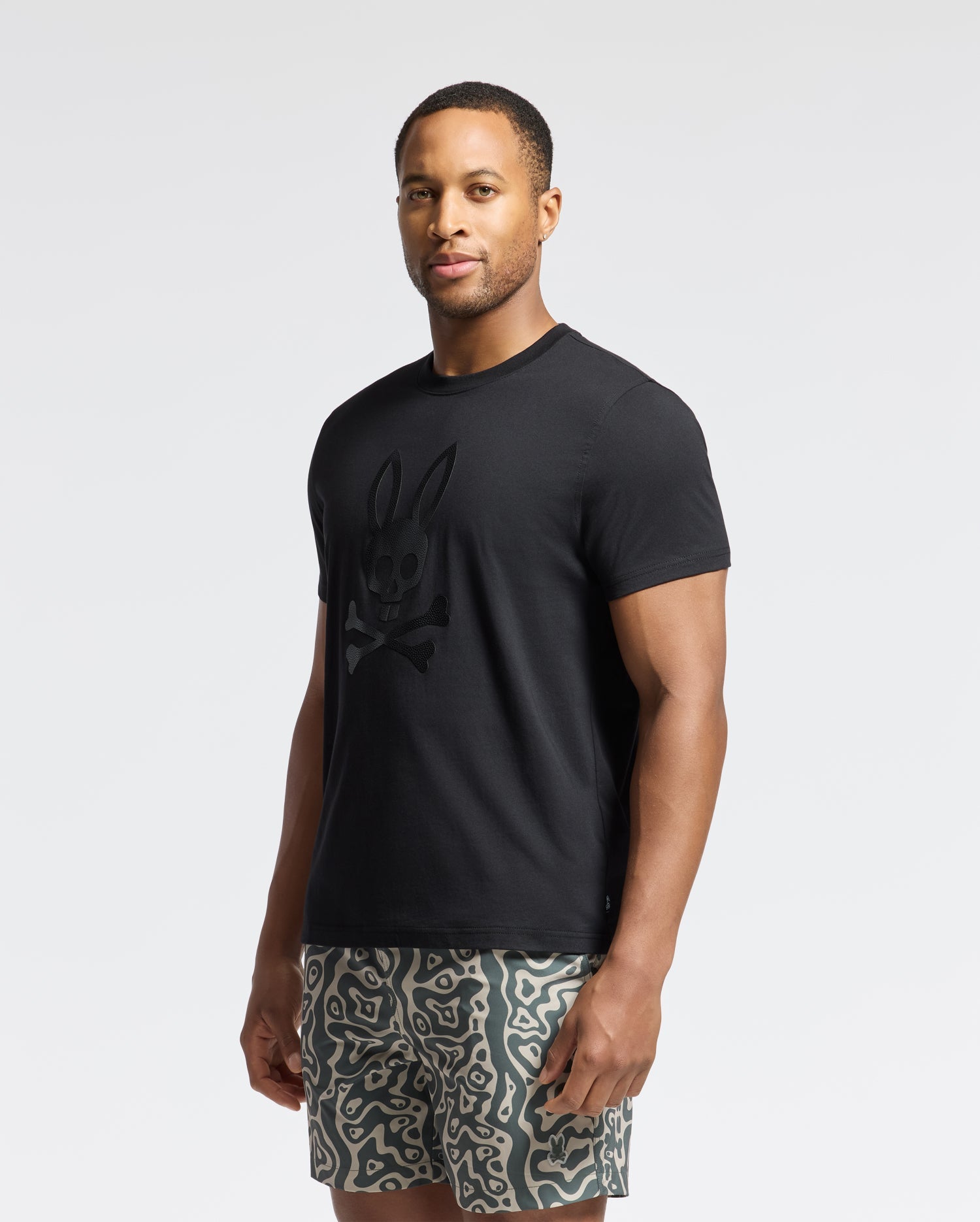 A man stands slightly to the side wearing a Psycho Bunny Mens Payson Leather Print Graphic Tee made from Pima cotton jersey, showcasing a rabbit skull and crossbones design with patterned shorts. The background is plain light gray.
