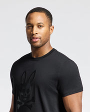 A person is wearing the Psycho Bunny MENS PAYSON LEATHER PRINT GRAPHIC TEE in black, crafted from Pima cotton jersey, and featuring a stylized rabbit skull and crossbones logo against a neutral background.
