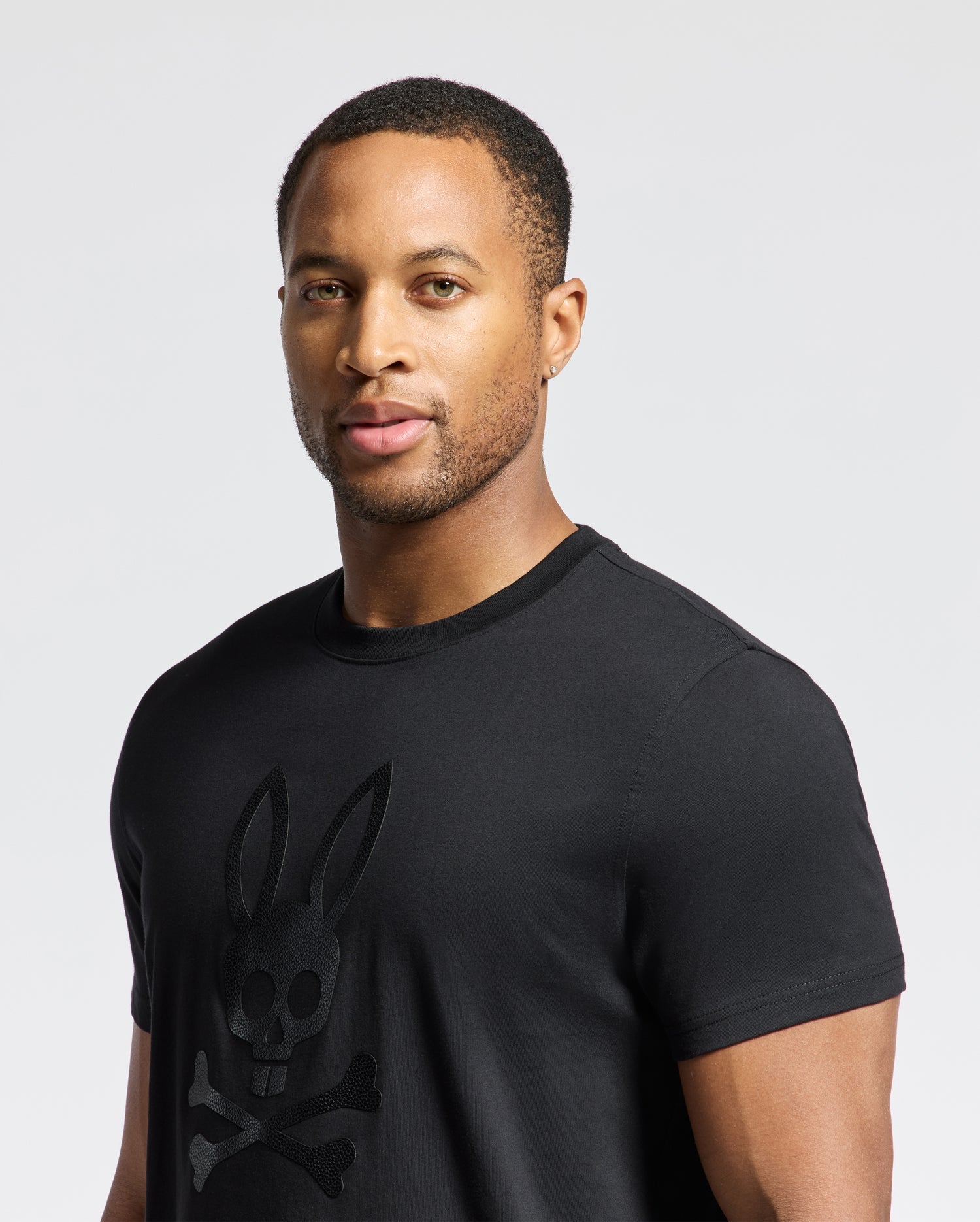 A person is wearing the Psycho Bunny MENS PAYSON LEATHER PRINT GRAPHIC TEE in black, crafted from Pima cotton jersey, and featuring a stylized rabbit skull and crossbones logo against a neutral background.