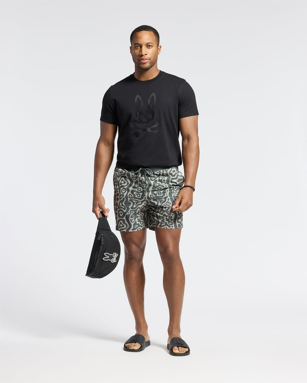A man wearing the MENS PAYSON LEATHER PRINT GRAPHIC TEE by Psycho Bunny, paired with patterned shorts and black slide sandals. He carries a matching black leather print bag against a plain white background.