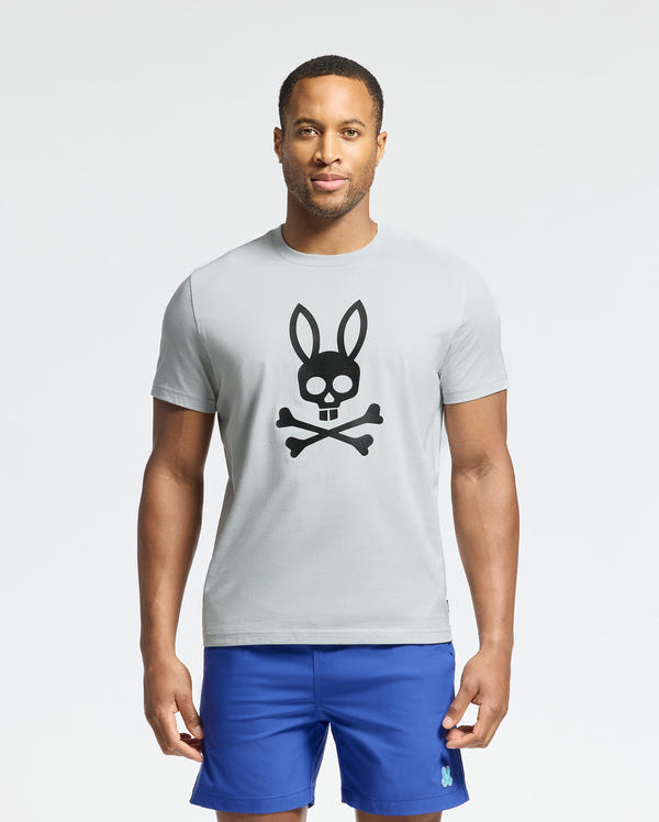 Against a plain background, someone wears the MENS PAYSON LEATHER PRINT GRAPHIC TEE from Psycho Bunny, light gray with a black bunny and crossbones design, paired with blue shorts.