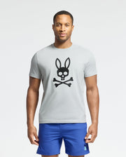 Against a plain background, someone wears the MENS PAYSON LEATHER PRINT GRAPHIC TEE from Psycho Bunny, light gray with a black bunny and crossbones design, paired with blue shorts.