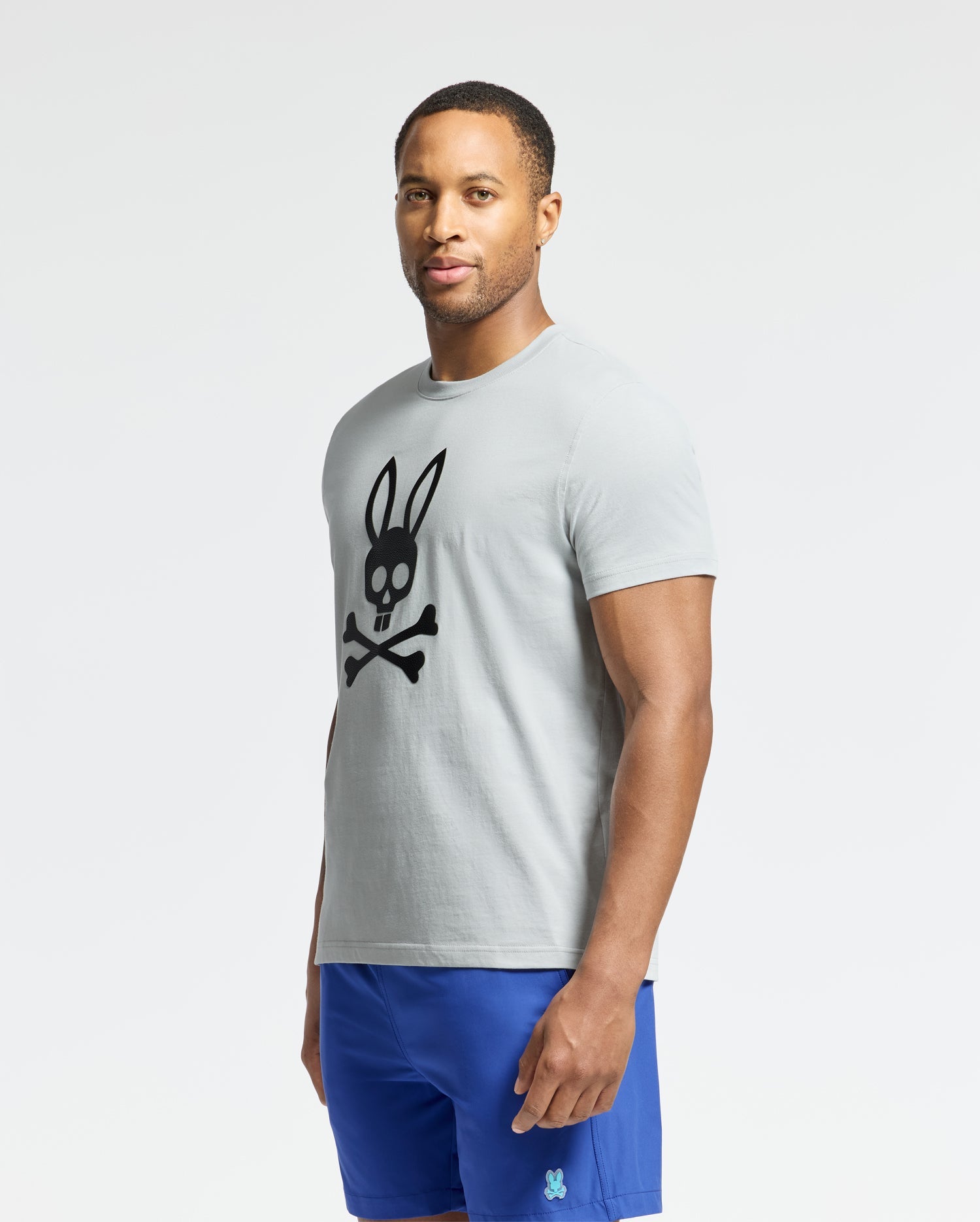 A man wears the Psycho Bunny MENS PAYSON LEATHER PRINT GRAPHIC TEE, featuring a black bunny skull and crossed bones graphic, along with bright blue shorts against a plain white background.
