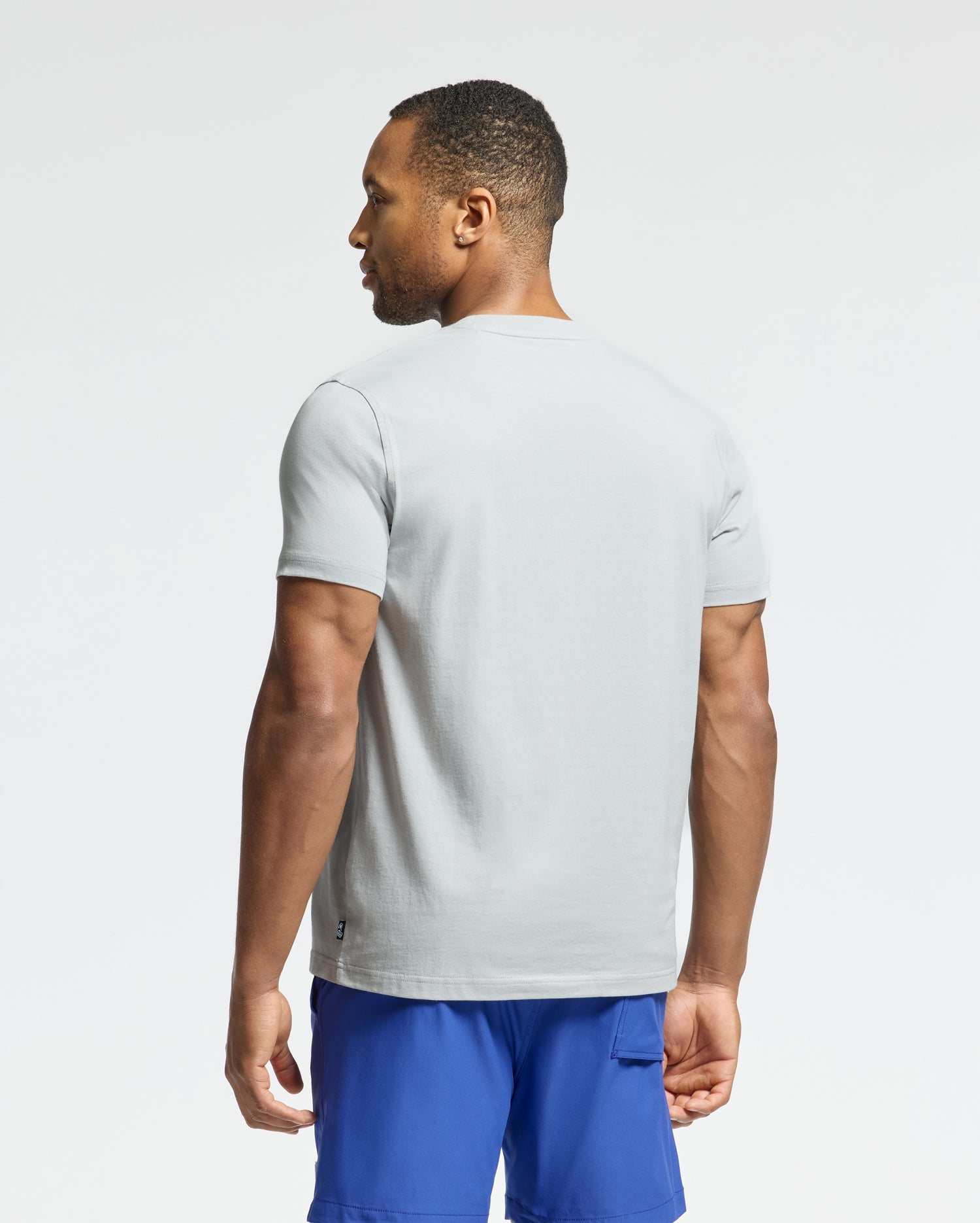 A man wearing the Psycho Bunny MENS PAYSON LEATHER PRINT GRAPHIC TEE is standing in light gray Pima cotton with blue shorts, facing away from the camera against a plain white background.