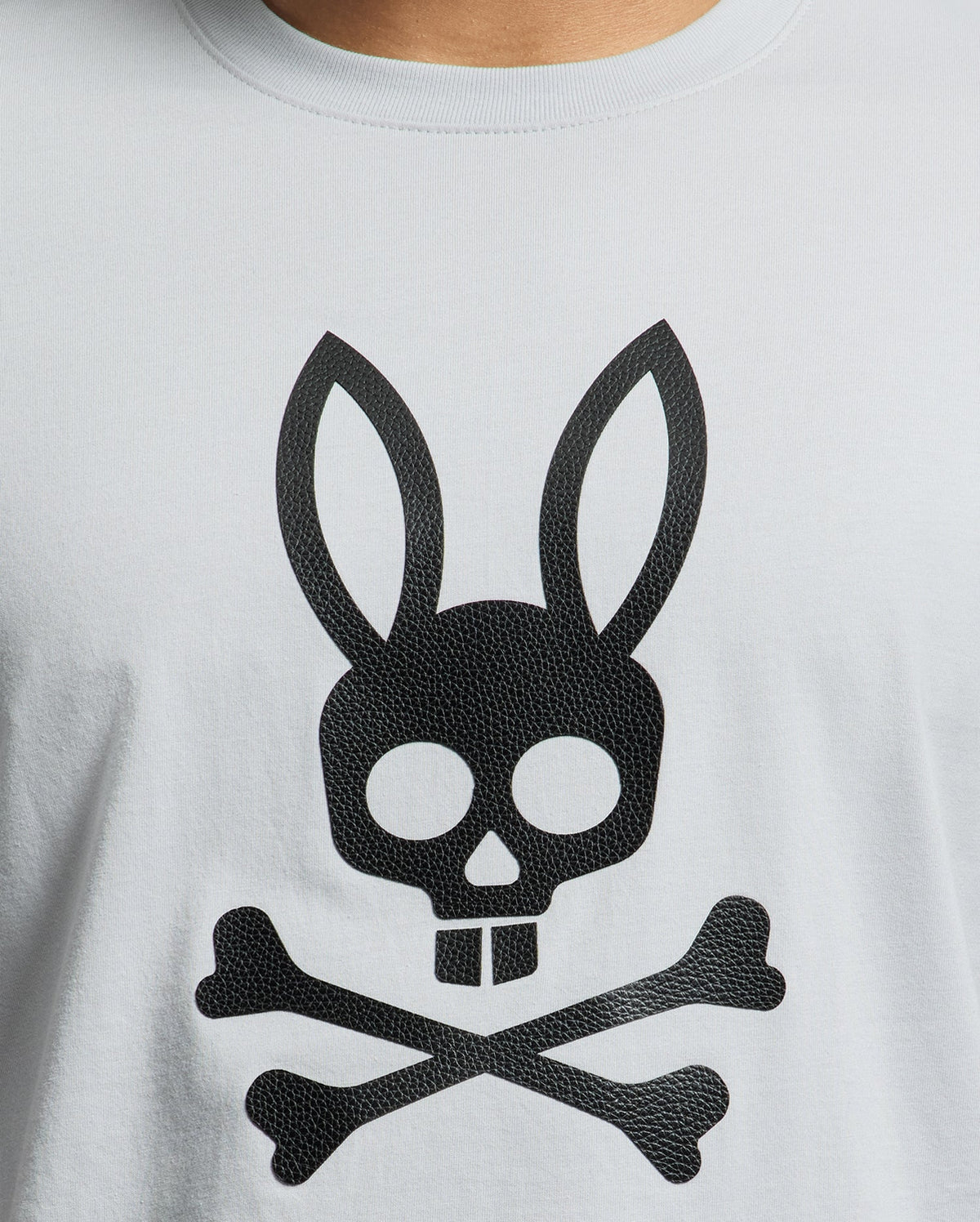 The Psycho Bunny MENS PAYSON LEATHER PRINT GRAPHIC TEE features a distinctive black skull with bunny ears and buck teeth, set above crossed bones on light gray Pima cotton. The leather print adds an edgy flair to this standout piece.