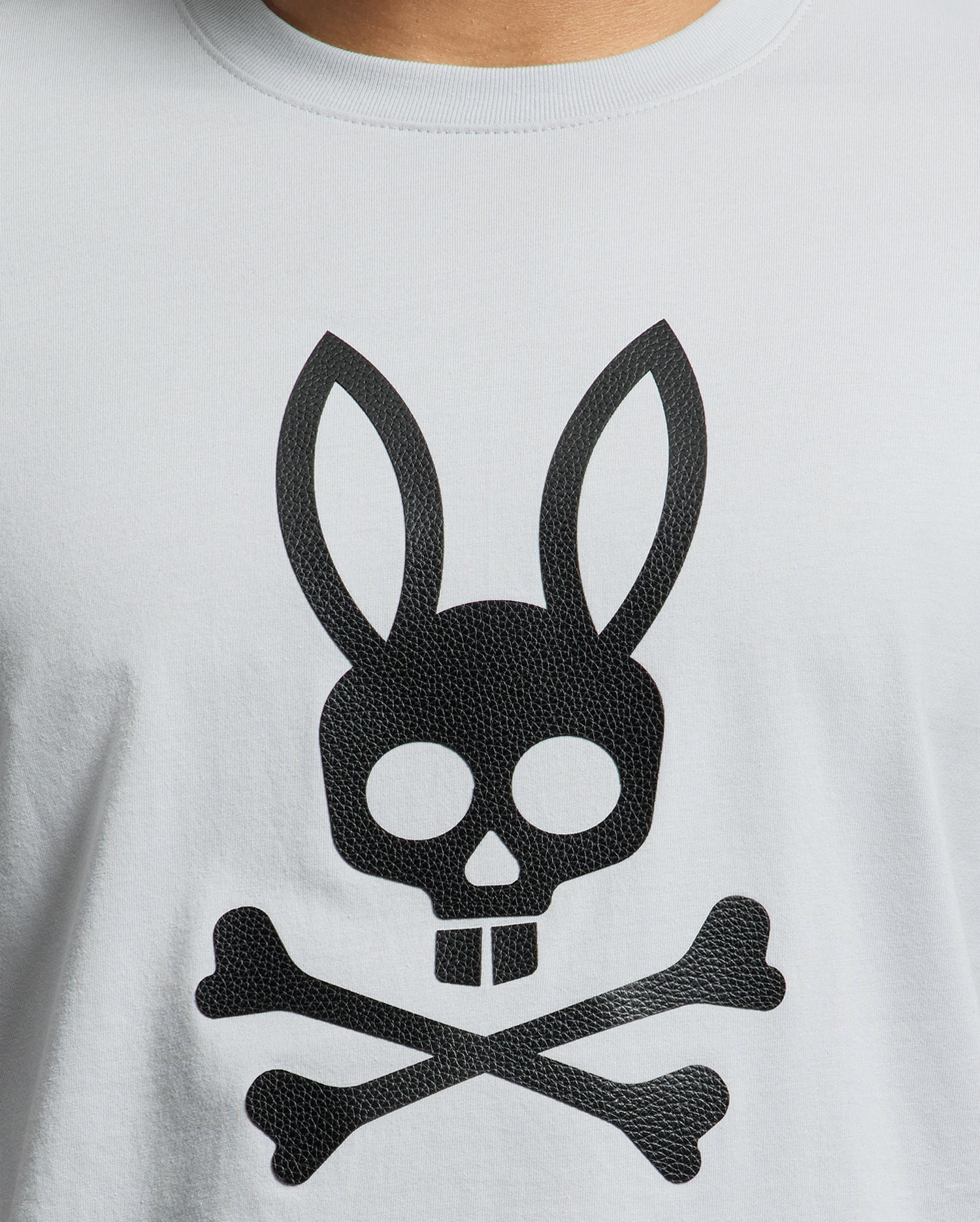 The Psycho Bunny MENS PAYSON LEATHER PRINT GRAPHIC TEE features a distinctive black skull with bunny ears and buck teeth, set above crossed bones on light gray Pima cotton. The leather print adds an edgy flair to this standout piece.