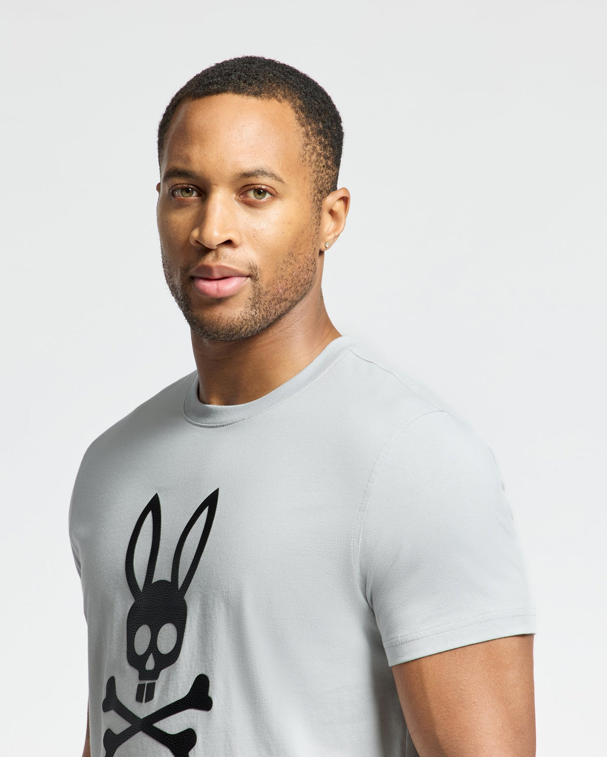 A person with short hair is wearing the Psycho Bunny MENS PAYSON LEATHER PRINT GRAPHIC TEE, made from Pima cotton in light gray, featuring a black skull with rabbit ears and crossbones. The background is plain white.