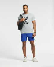 A man stands against a plain background wearing a light gray Psycho Bunny Payson Leather Print Graphic Tee, blue shorts, white sneakers, and a watch. He is carrying a black shoulder bag with a black bunny skull graphic.