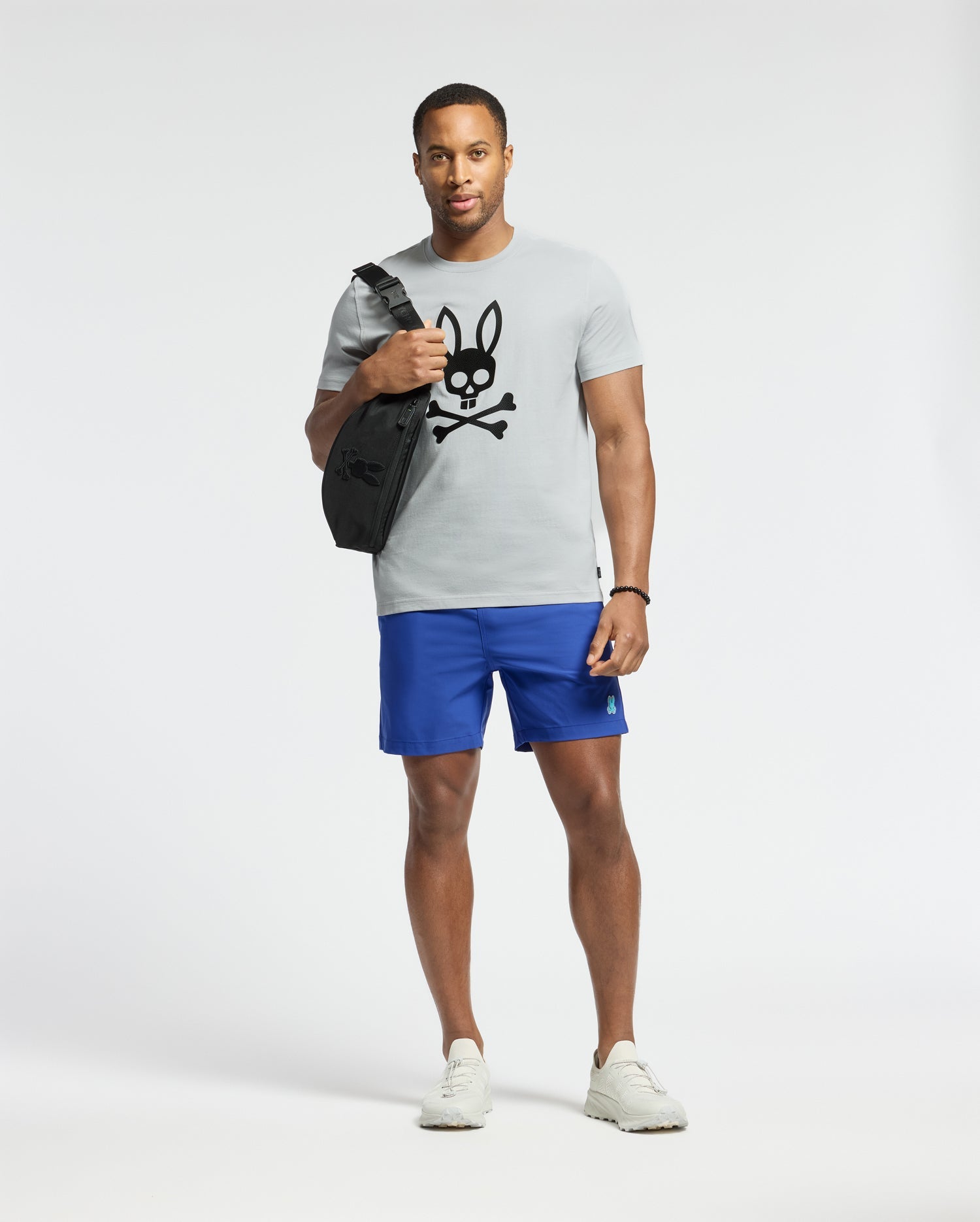 A man stands against a plain background wearing a light gray Psycho Bunny Payson Leather Print Graphic Tee, blue shorts, white sneakers, and a watch. He is carrying a black shoulder bag with a black bunny skull graphic.