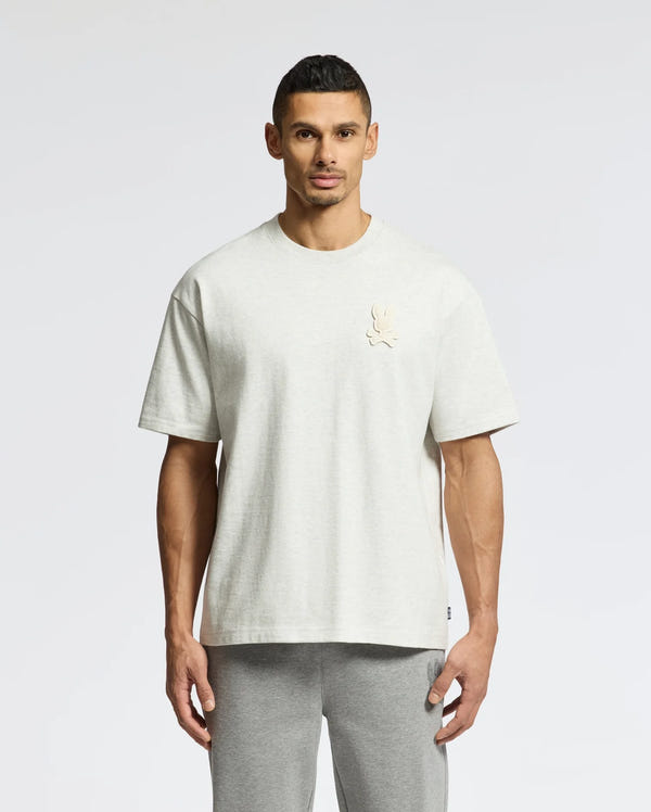 A person in a Psycho Bunny MENS ELDORADO OVERSIZED TEE, light gray, made from relaxed-fit heavyweight Pima cotton jersey with a chest emblem, stands against a plain white background. They're also wearing matching gray pants.