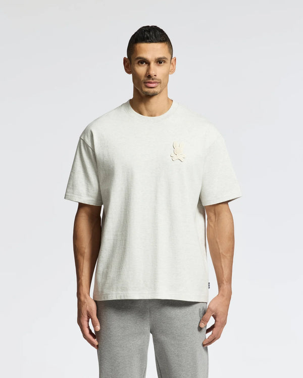 A person in a Psycho Bunny MENS ELDORADO OVERSIZED TEE, light gray, made from relaxed-fit heavyweight Pima cotton jersey with a chest emblem, stands against a plain white background. They're also wearing matching gray pants.