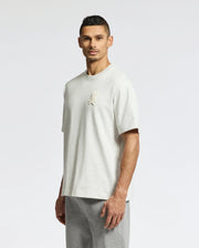 A man in a relaxed fit, light gray Psycho Bunny MENS ELDORADO OVERSIZED TEE - B6U215E200, made from heavyweight Pima cotton jersey with a small embroidered chest design, pairs with gray pants against a plain white backdrop.