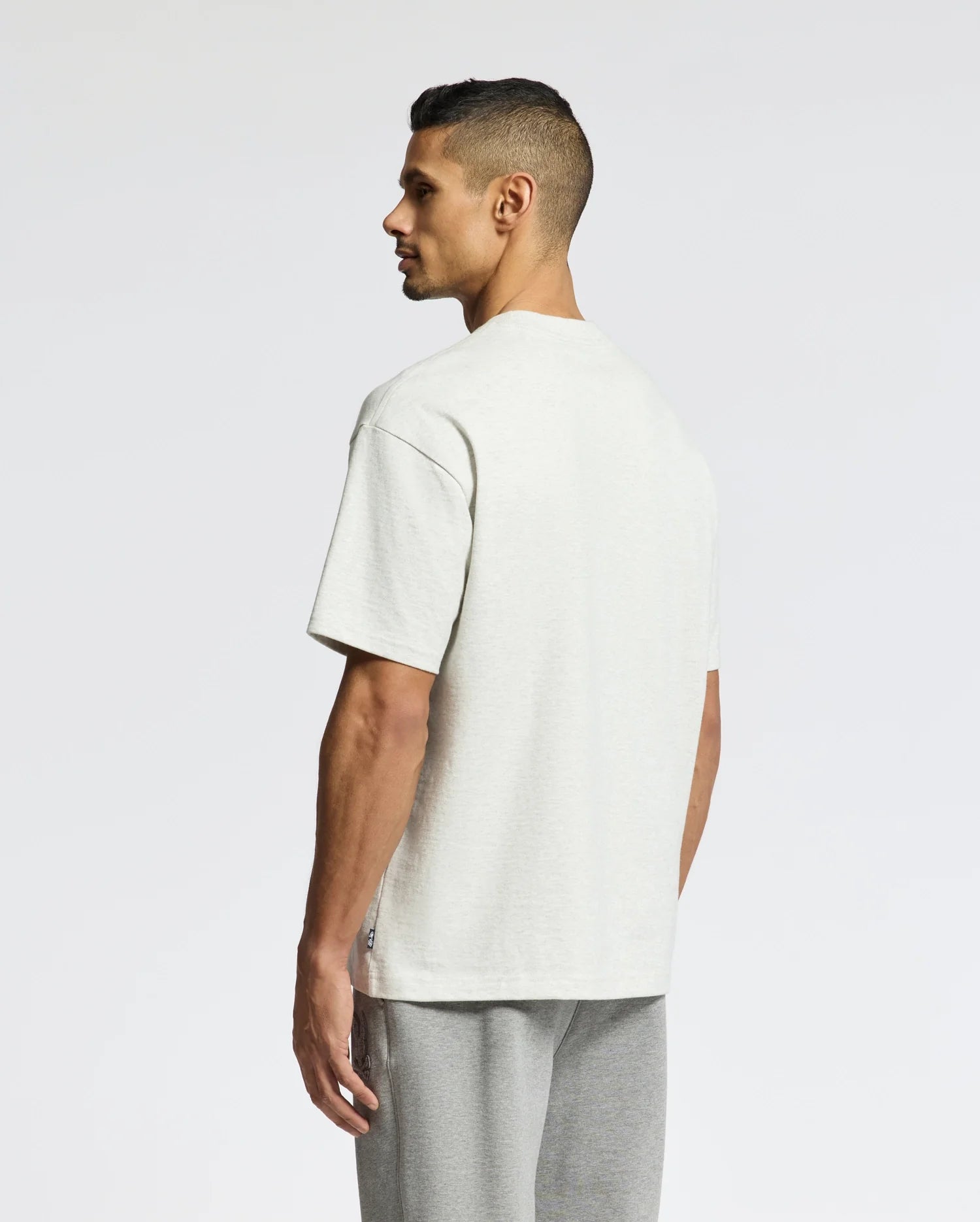 A person with short dark hair stands sideways, looking away. They're wearing a Psycho Bunny MENS ELDORADO OVERSIZED TEE - B6U215E200 in light gray, made from heavyweight Pima cotton jersey, paired with matching gray pants against a plain white background.