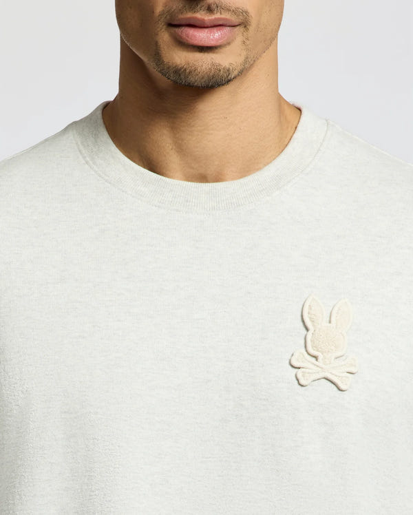 Wearing the Psycho Bunny "MENS ELDORADO OVERSIZED TEE - B6U215E200", a relaxed-fit, light gray heavyweight Pima cotton jersey features an embroidered beige cartoon bunny head with crossbones on the left chest against a plain neutral background.