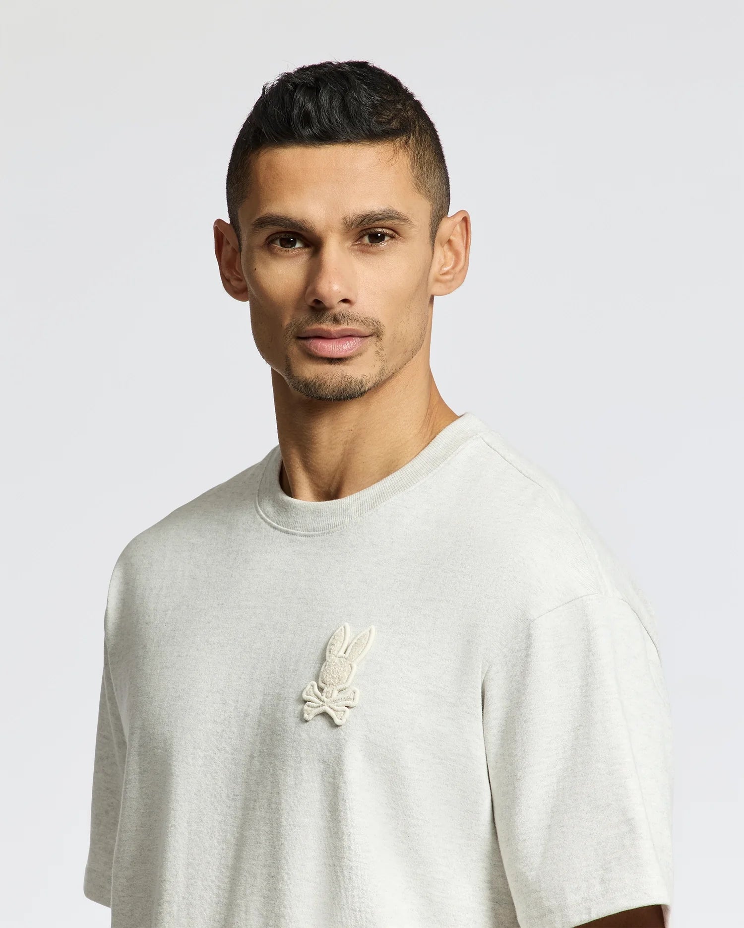 A man with short dark hair and a trimmed beard is wearing the Psycho Bunny MENS ELDORADO OVERSIZED TEE - B6U215E200, crafted from heavyweight Pima cotton jersey with an embroidered bunny and crossbones on the chest. The relaxed fit drapes comfortably as he stands against a plain background.