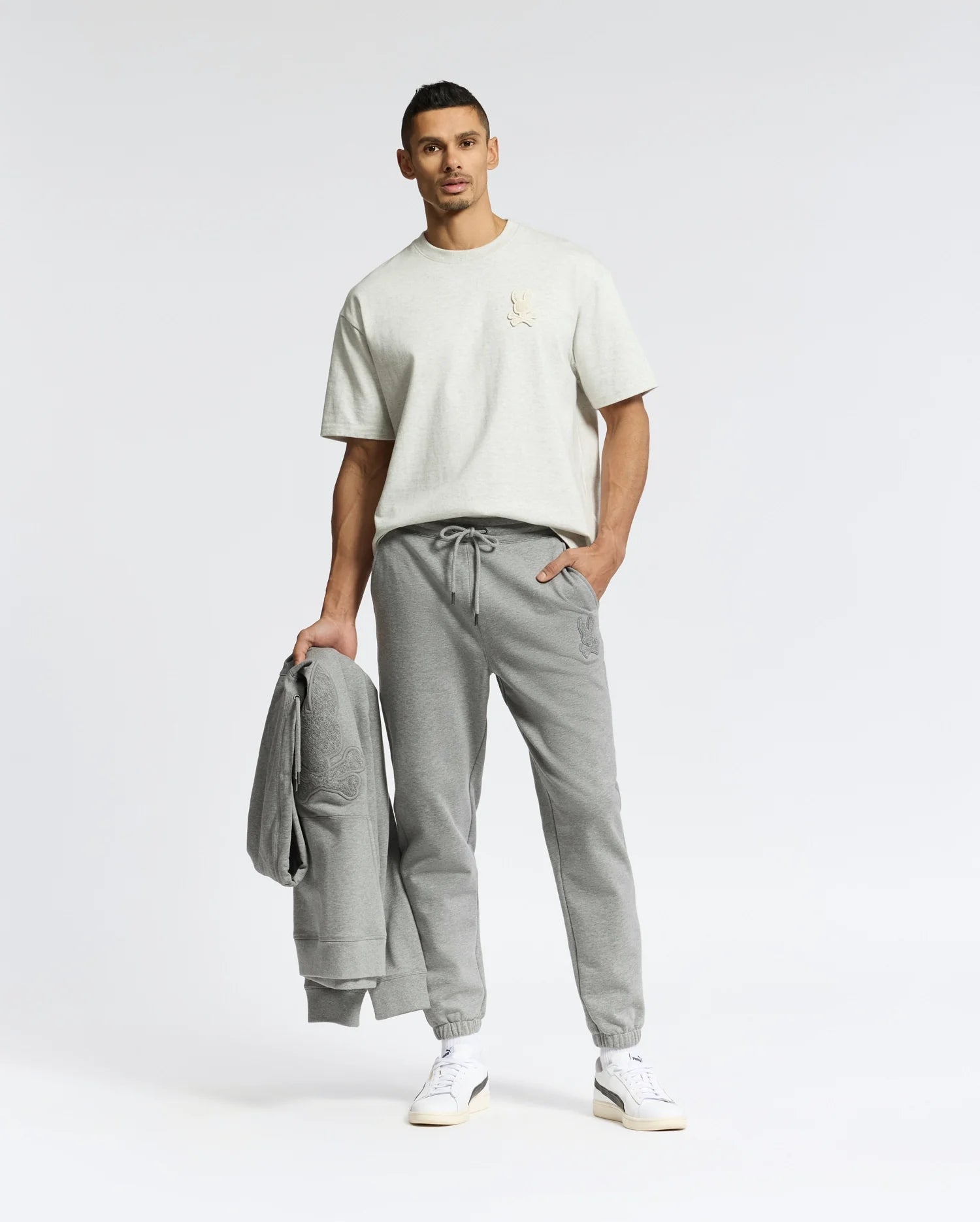 A man stands against a plain backdrop wearing Psycho Bunny's MENS ELDORADO OVERSIZED TEE in beige, crafted from heavyweight Peruvian Pima cotton, with relaxed-fit grey joggers and white sneakers. He holds a matching grey sweatshirt in his left hand, while his right hand is in his pocket.