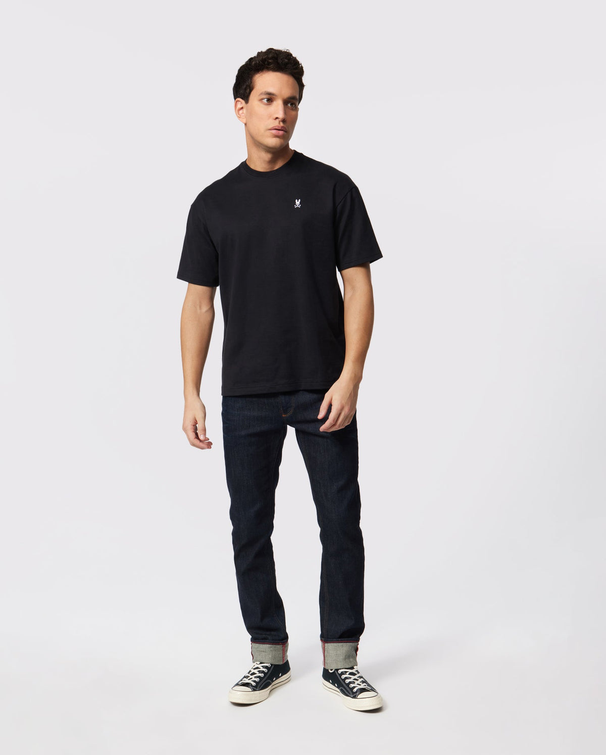 A person with short, dark hair stands against a plain white background. They are wearing a black t-shirt featuring an embroidered bunny logo, dark blue jeans, and black and white sneakers. Their posture is relaxed with hands by their sides, exuding casual confidence in their MENS OVERSIZED CLASSIC CREW NECK TEE - B6U240W1PC by Psycho Bunny.