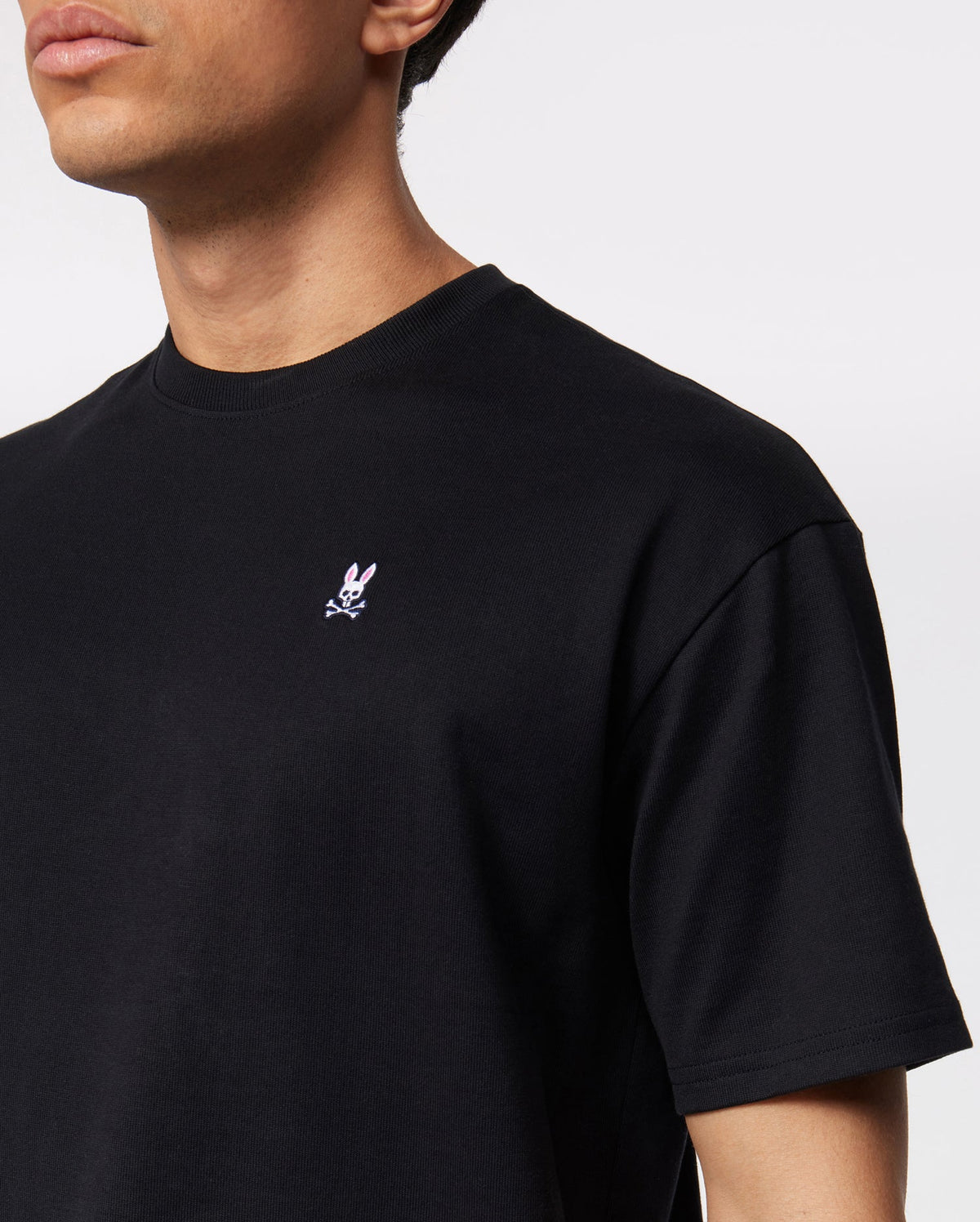 A person wearing a Psycho Bunny MENS OVERSIZED CLASSIC CREW NECK TEE - B6U240W1PC black t-shirt with a small white and pink embroidered bunny logo on the upper left chest. The background is plain white.