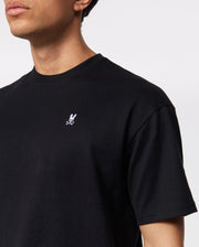 A person wearing a Psycho Bunny MENS OVERSIZED CLASSIC CREW NECK TEE - B6U240W1PC black t-shirt with a small white and pink embroidered bunny logo on the upper left chest. The background is plain white.