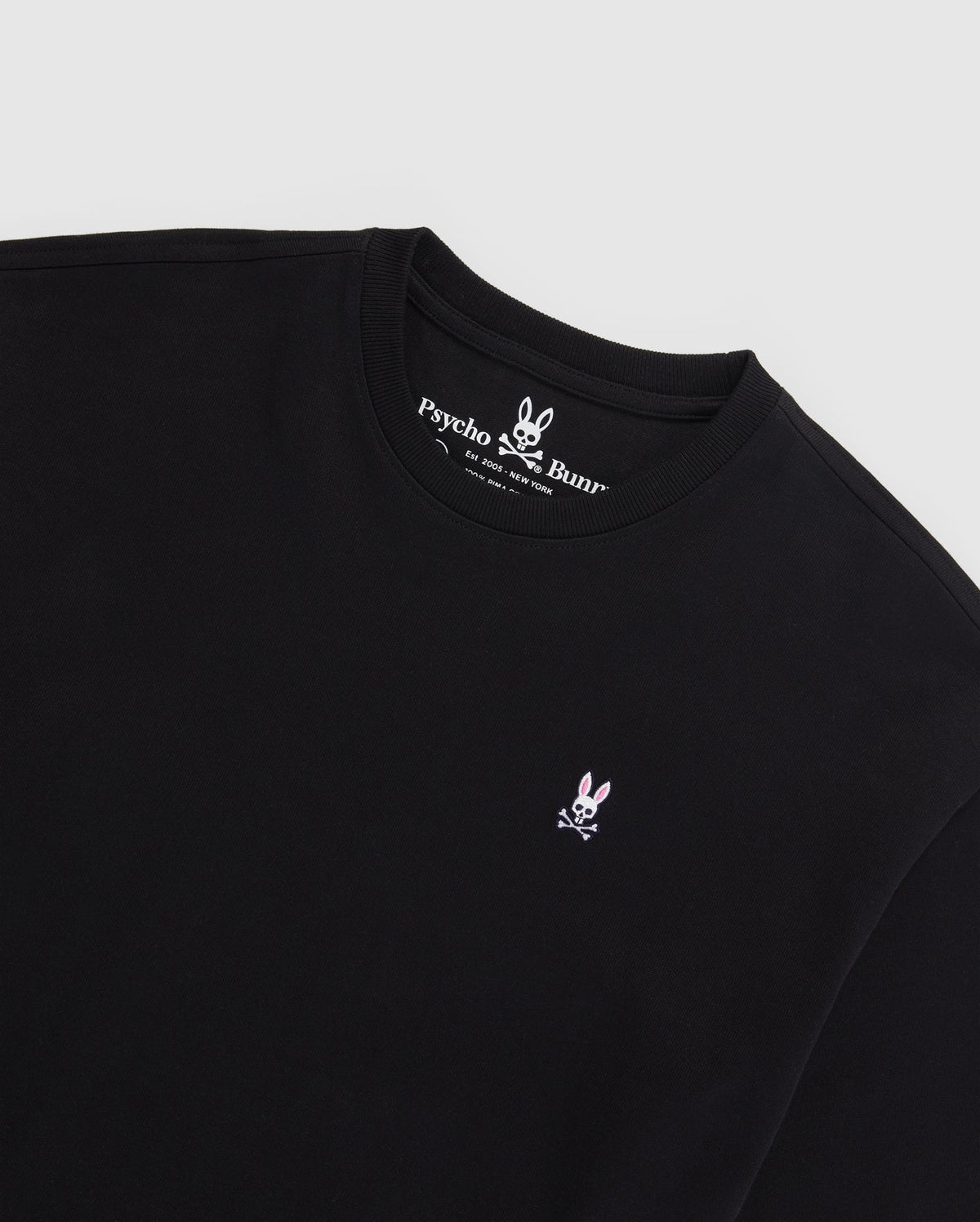 A black MENS OVERSIZED CLASSIC CREW NECK TEE - B6U240W1PC from Psycho Bunny, featuring the brand's Embroidered Bunny logo—a white bunny with crossed bones beneath it—stitched in pink and white on the left chest. Made from luxurious Peruvian Pima cotton, the inner collar showcases the brand name and logo in white text on a black background.