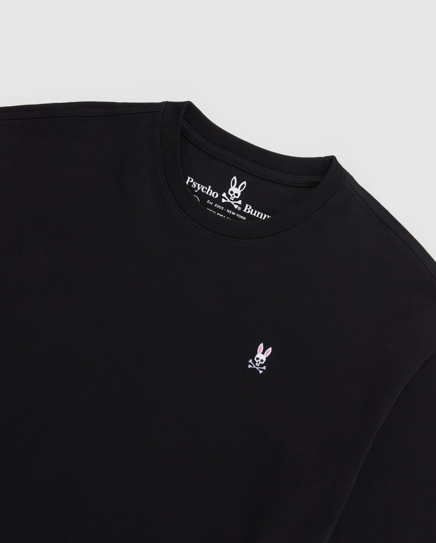 A black MENS OVERSIZED CLASSIC CREW NECK TEE - B6U240W1PC from Psycho Bunny, featuring the brand's Embroidered Bunny logo—a white bunny with crossed bones beneath it—stitched in pink and white on the left chest. Made from luxurious Peruvian Pima cotton, the inner collar showcases the brand name and logo in white text on a black background.