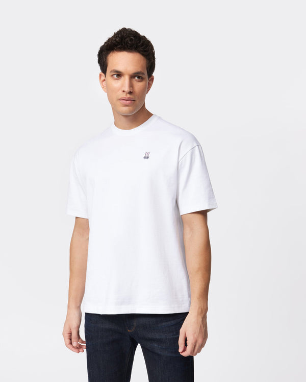 A man with short dark hair is wearing a plain white MENS OVERSIZED CLASSIC CREW NECK TEE - B6U240W1PC from Psycho Bunny and dark jeans, standing against a plain white background. The heavyweight fit t-shirt has a small embroidered logo on the left side of the chest. The man is looking slightly to the side with a neutral expression.