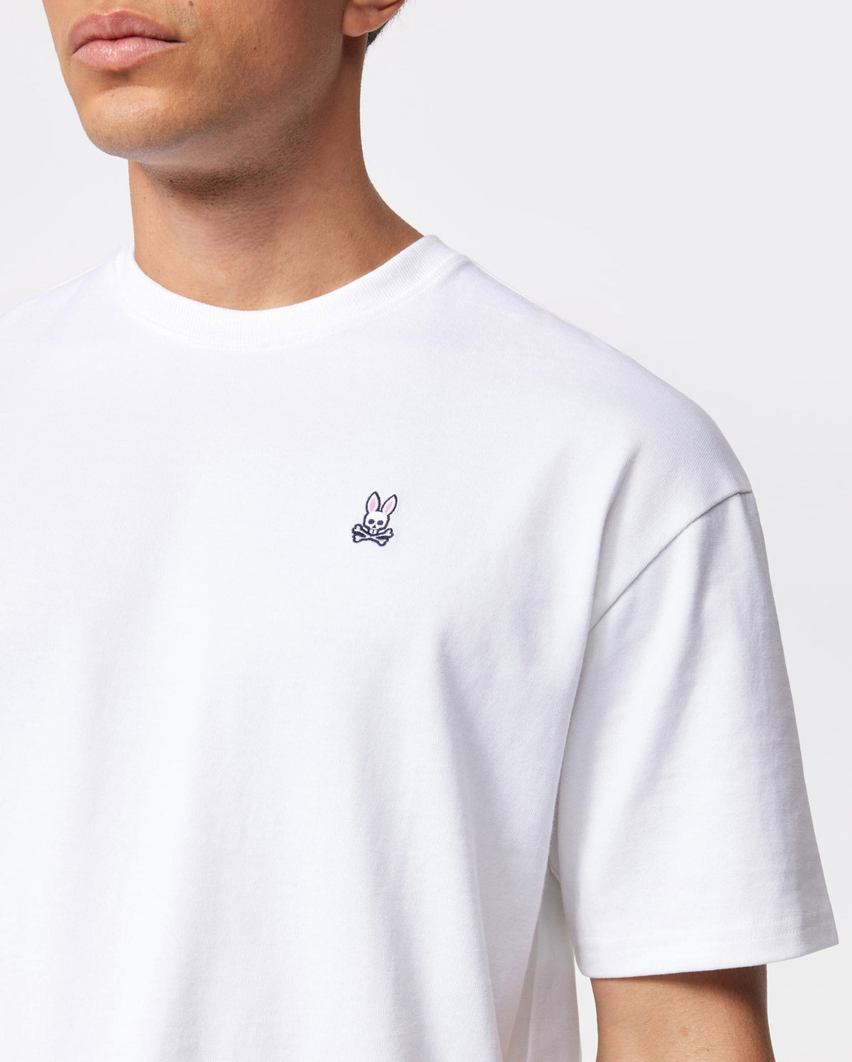 A man wearing a Psycho Bunny MENS OVERSIZED CLASSIC CREW NECK TEE - B6U240W1PC with a small embroidered pink bunny and crossbones emblem on the left chest. The background is plain white. The man is shown from the shoulders up, with his face partially out of frame.