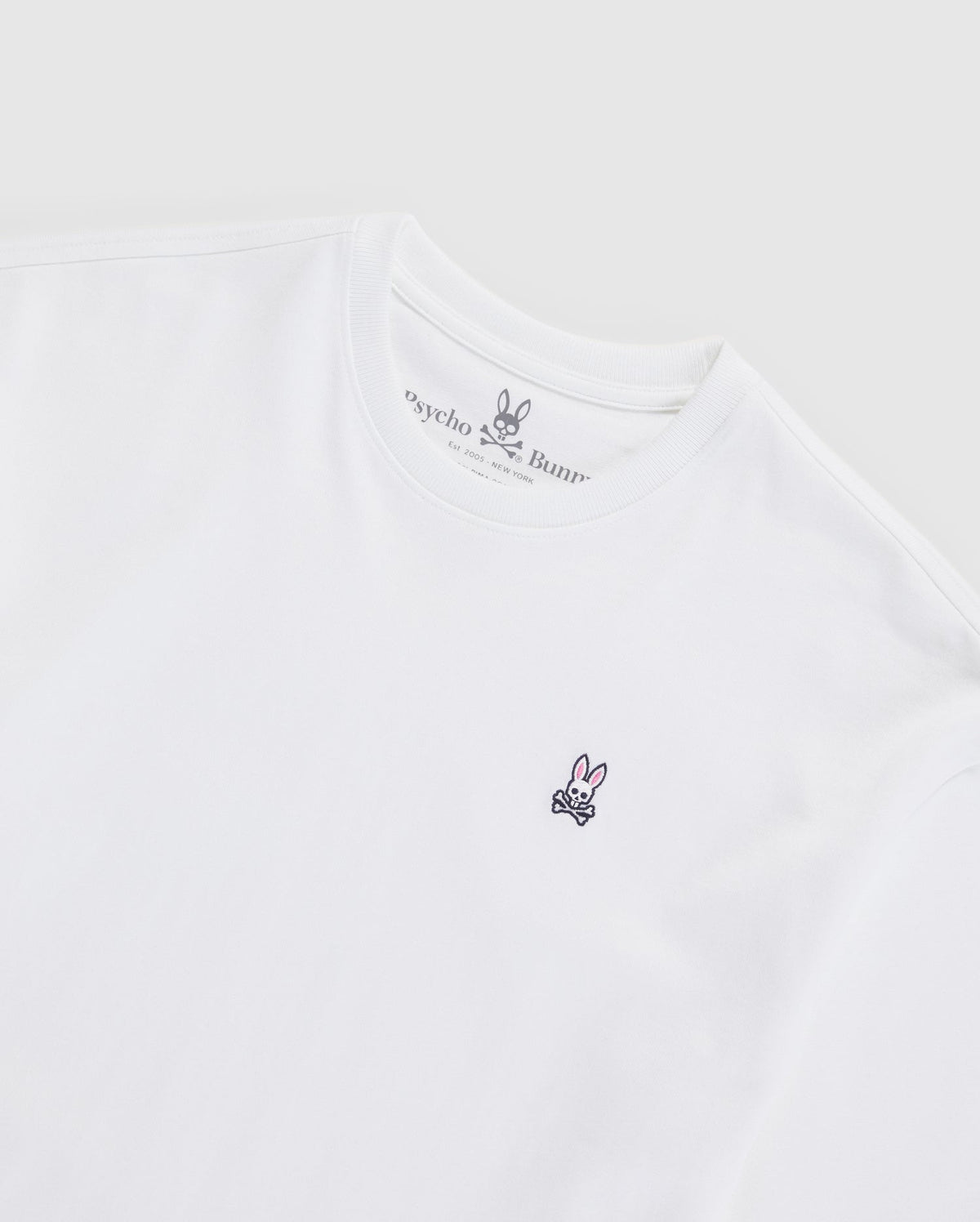 A white MENS OVERSIZED CLASSIC CREW NECK TEE - B6U240W1PC with a minimalist design featuring a small embroidered logo of a bunny with crossed bones below it on the left chest. Made from soft Pima Cotton, it has a round crew neck and offers a smooth, heavyweight fit. Brand Name: Psycho Bunny.