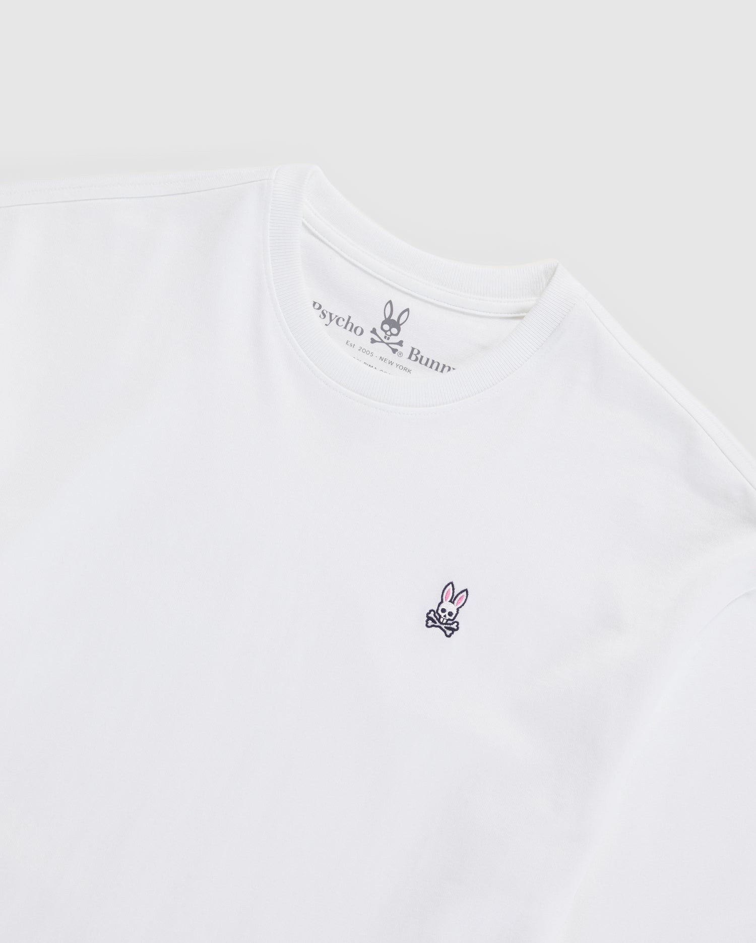 A white MENS OVERSIZED CLASSIC CREW NECK TEE - B6U240W1PC with a minimalist design featuring a small embroidered logo of a bunny with crossed bones below it on the left chest. Made from soft Pima Cotton, it has a round crew neck and offers a smooth, heavyweight fit. Brand Name: Psycho Bunny.