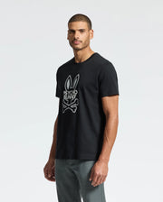 A man wearing a black MENS DUMON GRAPHIC TEE - B6U271E200 featuring the "Psycho Bunny" logo stands against a light gray background. Made from Premium Pima Cotton, he sports short hair and a trimmed beard, paired with gray pants. He is facing slightly to the left with one arm by his side.