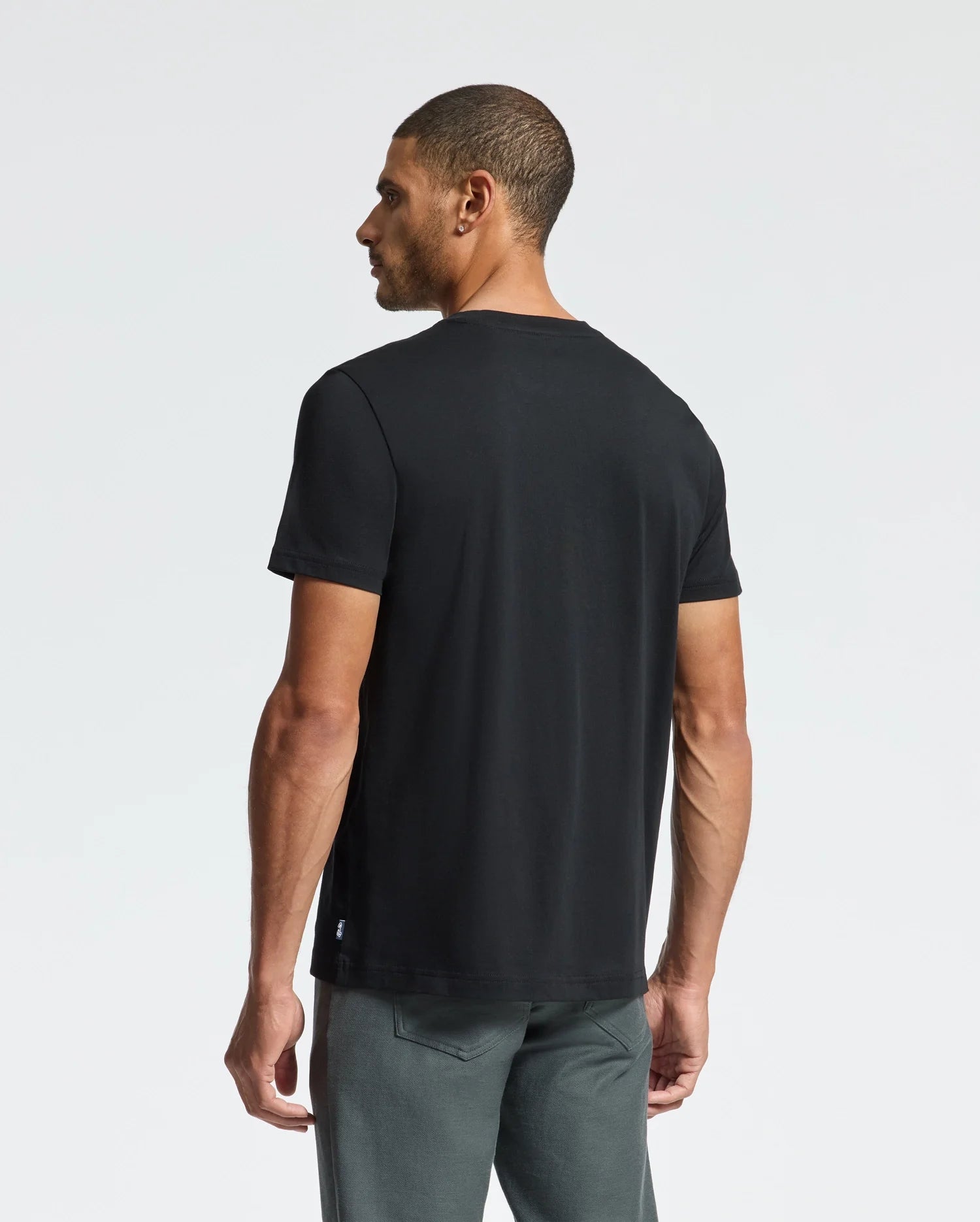 A man in a Psycho Bunny MENS DUMON GRAPHIC TEE - B6U271E200, crafted from premium Pima cotton, and gray pants stands with his back turned, glancing to the side against a white minimalist background.